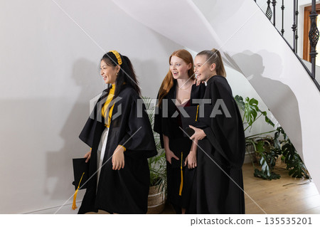 Teen graduates in caps and gowns celebrating together at home, smiling joyfully 135535201