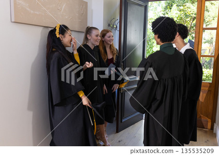 Teens in graduation gowns celebrating together at home, smiling and chatting Teens in graduation gowns celebrating together at home, smiling and chatting 135535209