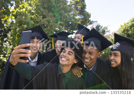 Graduating students in caps and gowns taking selfie, celebrating outdoors together Graduating students in caps and gowns taking selfie, celebrating outdoors together 135535218