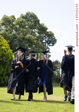 Graduating students in caps and gowns walking joyfully on grassy field Graduating students in caps and gowns walking joyfully on grassy field 135535233