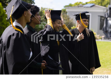 Graduating students adjusting caps and smiling joyfully while celebrating outdoors 135535238