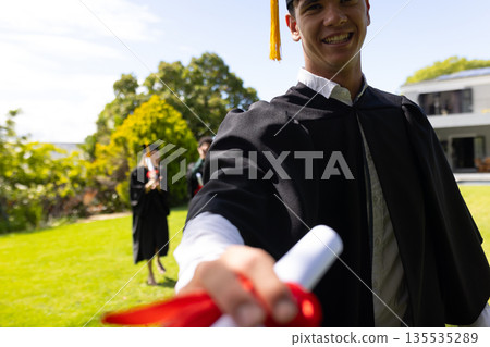Holding diploma, graduate smiling outdoors celebrating academic achievement with friends Holding diploma, graduate smiling outdoors celebrating academic achievement with friends 135535289