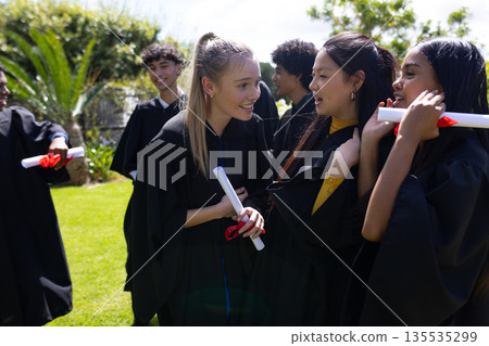 Celebrating outdoors, teens in graduation gowns holding diplomas, smiling joyfully 135535299