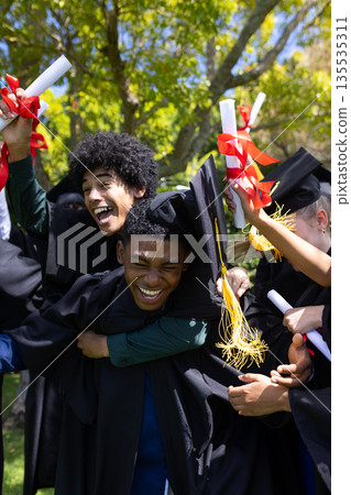 Graduating students in caps and gowns celebrating outdoors, holding diplomas 135535311