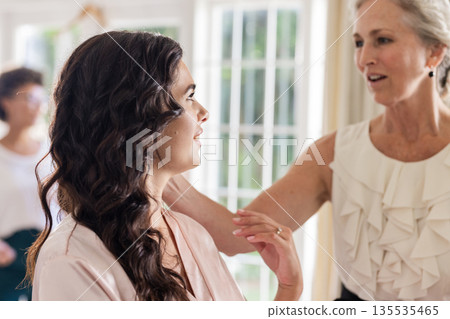 Bride getting ready, smiling with woman helping adjust hair in bright room Bride getting ready, smiling with woman helping adjust hair in bright room 135535465
