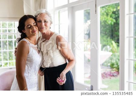 Embracing older woman, bride smiling warmly in sunlit room, celebrating together 135535485