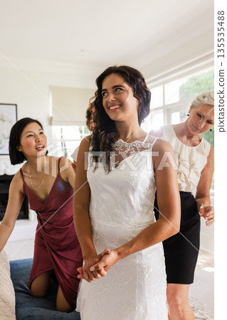 Bride in elegant white dress smiling with bridesmaids helping at home 135535488