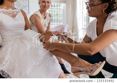Bride wearing lace garter while friends assist, preparing for wedding ceremony Bride wearing lace garter while friends assist, preparing for wedding ceremony 135535492