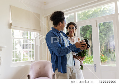 Bride holding bouquet smiling at groom in bright living room, sharing joyful moment Bride holding bouquet smiling at groom in bright living room, sharing joyful moment 135535500