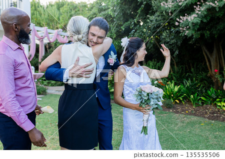 Groom hugging guest warmly while bride holding bouquet, smiling in garden 135535506