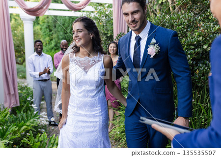 Bride and groom walking joyfully down garden aisle, guests applauding happily 135535507