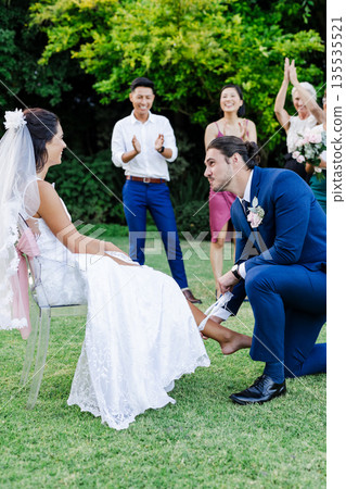 Groom kneeling, removing garter from bride's leg, friends cheering in garden 135535521