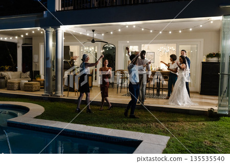 Celebrating joyfully, guests at outdoor wedding reception near pool with string lights 135535540