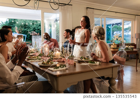 Bride giving speech at elegant dinner table, guests applauding and smiling warmly 135535543