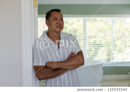 Senior man standing by window at home, reflecting with arms crossed Senior man standing by window at home, reflecting with arms crossed 135535544