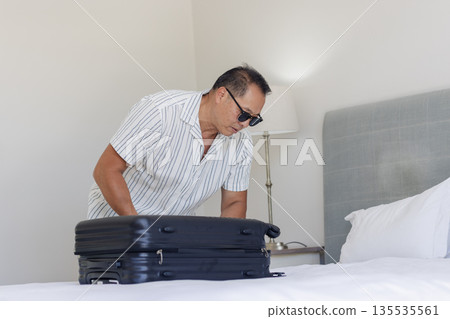 Senior man packing suitcase on bed, preparing for travel with sunglasses on Senior man packing suitcase on bed, preparing for travel with sunglasses on 135535561