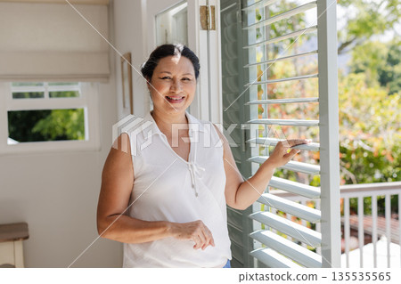 Smiling woman enjoying sunlight at home, standing by open window shutters Smiling woman enjoying sunlight at home, standing by open window shutters 135535565
