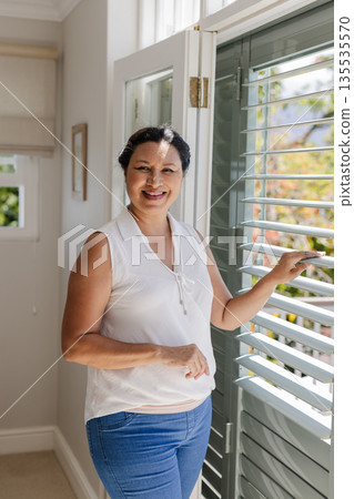 Senior woman smiling while opening window blinds at home, enjoying sunlight Senior woman smiling while opening window blinds at home, enjoying sunlight 135535570