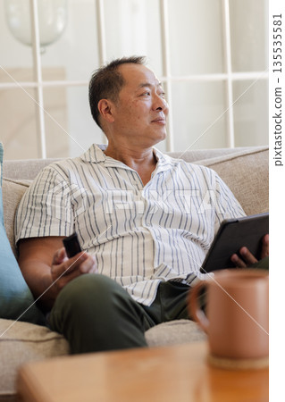 Senior man relaxing on sofa holding tablet and remote, enjoying leisure time Senior man relaxing on sofa holding tablet and remote, enjoying leisure time 135535581
