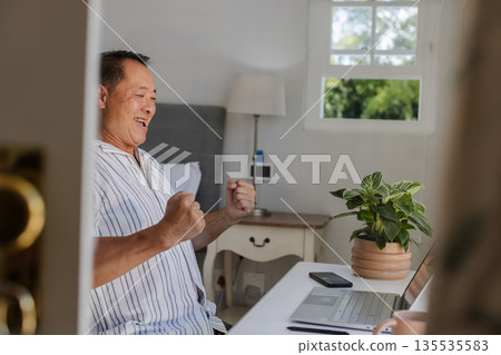 Senior man celebrating success while using laptop at home office desk Senior man celebrating success while using laptop at home office desk 135535583