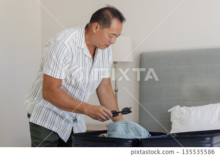 Senior man packing suitcase at home, preparing for travel with sunglasses Senior man packing suitcase at home, preparing for travel with sunglasses 135535586