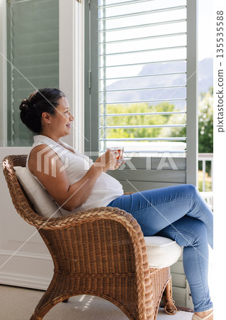 Relaxing at home, woman enjoying coffee while sitting in wicker chair by window 135535588