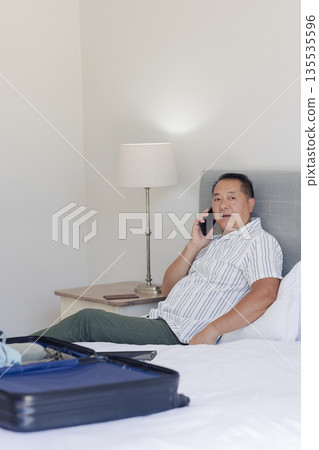Senior man relaxing on bed, talking on smartphone with suitcase nearby, at home Senior man relaxing on bed, talking on smartphone with suitcase nearby, at home 135535596