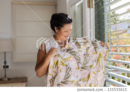 Asian woman happily holding floral shirt near window at home Asian woman happily holding floral shirt near window at home 135535615