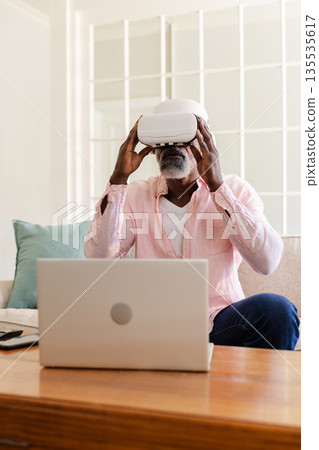 Senior man using VR headset while sitting on sofa with laptop nearby Senior man using VR headset while sitting on sofa with laptop nearby 135535617