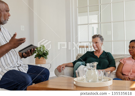 African American man discussing with senior women on couch at home, holding tablet African American man discussing with senior women on couch at home, holding tablet 135535618