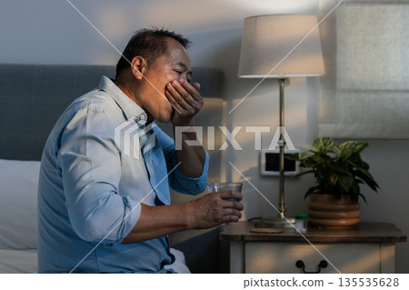 Asian man yawning while holding glass of water in bedroom, looking tired Asian man yawning while holding glass of water in bedroom, looking tired 135535628