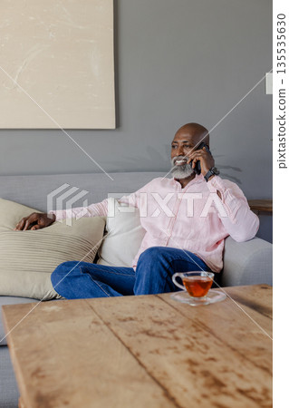 Senior man in pink shirt relaxing on sofa, talking on smartphone with tea nearby 135535630