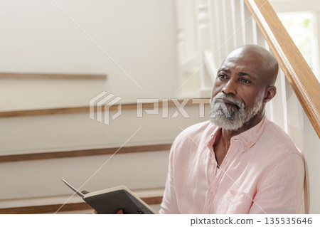 Elderly man sitting on stairs, reading book and reflecting peacefully at home 135535646