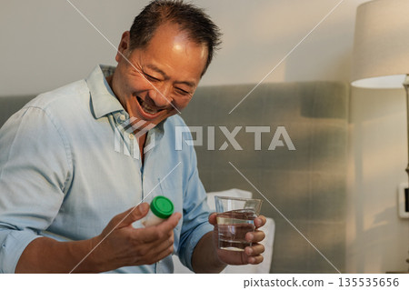Senior man joyfully holding medication bottle and glass of water at home 135535656