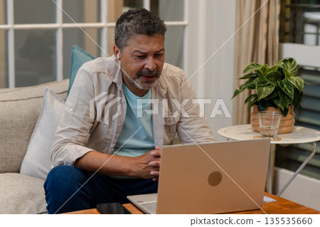 Senior man using laptop at home, wearing earbuds, focused on video call Senior man using laptop at home, wearing earbuds, focused on video call 135535660