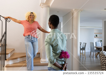 Smiling woman on stairs talking to man holding flowers behind back at home Smiling woman on stairs talking to man holding flowers behind back at home 135535724