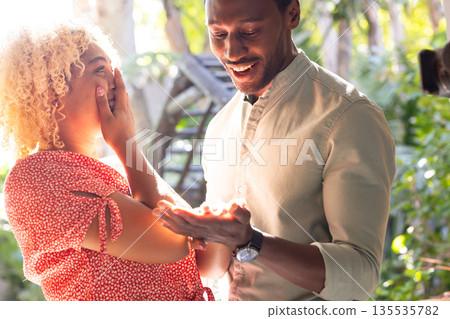 African American man surprising friend with gift outdoors, both looking delighted African American man surprising friend with gift outdoors, both looking delighted 135535782