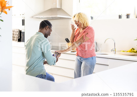 African American man proposing to surprised woman in bright kitchen, both smiling 135535791