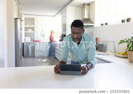 African American man using tablet in modern kitchen while friend cooks African American man using tablet in modern kitchen while friend cooks 135535801