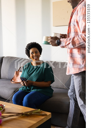 Smiling woman holding smartphone on couch while friend offers coffee mugs 135535839