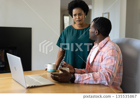 Man using smartphone and laptop at home, talking with concerned woman holding mug Man using smartphone and laptop at home, talking with concerned woman holding mug 135535867