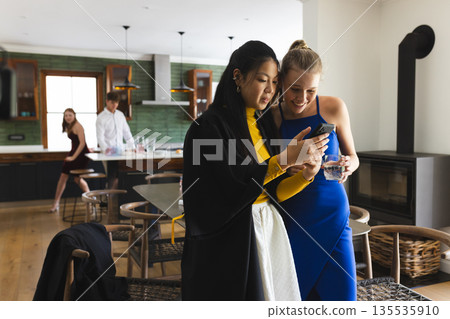 Teen girls in elegant dresses sharing smartphone moment at home party Teen girls in elegant dresses sharing smartphone moment at home party 135535910