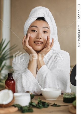 A young Asian woman with a towel on her head and wearing a bathrobe applies cream to her face, surrounded by skincare products and natural ingredients A young Asian woman with a towel on her head and wearing a bathrobe applies cream to her face, surrounded by skincare products and natural ingredients 135535925