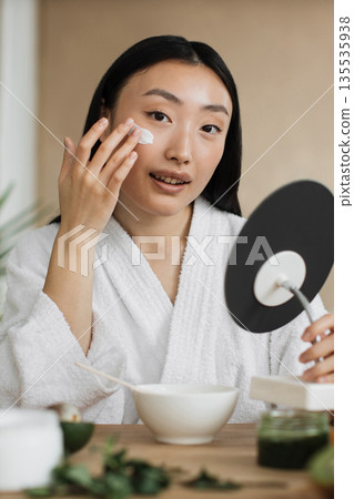 A young Asian woman in a white bathrobe applies cream to her face while looking in a mirror, surrounded by skincare ingredients 135535938