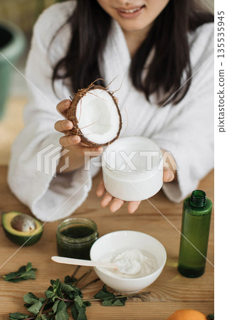 A woman in a white robe prepares natural skincare ingredients including coconut, avocado, and mint for a homemade beauty treatment A woman in a white robe prepares natural skincare ingredients including coconut, avocado, and mint for a homemade beauty treatment 135535945