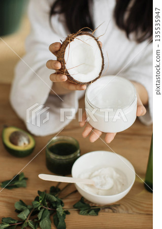 A person prepares a natural skincare product using fresh coconut, avocado, and mint on a wooden table 135535947