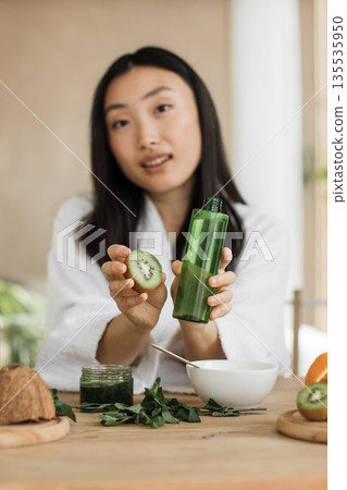 A young Asian woman in a white robe holds a kiwi slice and a green bottle of serum, surrounded by ingredients for homemade beauty products A young Asian woman in a white robe holds a kiwi slice and a green bottle of serum, surrounded by ingredients for homemade beauty products 135535950