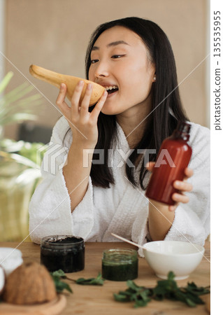 A young Asian woman in a white robe tastes a piece of butternut squash while holding a bottle of serum and surrounded by natural skincare ingredients 135535955