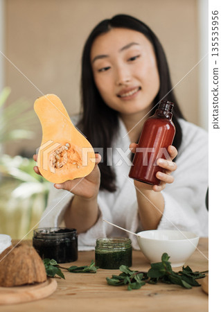 A young Asian woman in a white robe holds a halved butternut squash and a bottle of serum, surrounded by ingredients for natural skincare A young Asian woman in a white robe holds a halved butternut squash and a bottle of serum, surrounded by ingredients for natural skincare 135535956