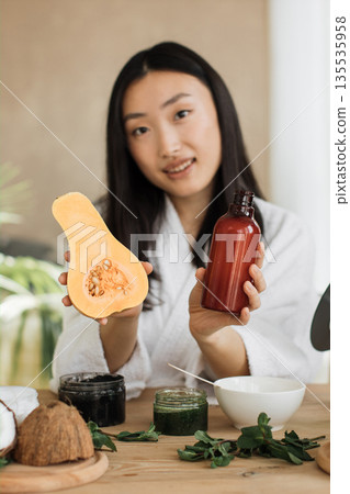A woman holds a butternut squash half and a bottle of cosmetic product, surrounded by ingredients for homemade beauty treatments 135535958